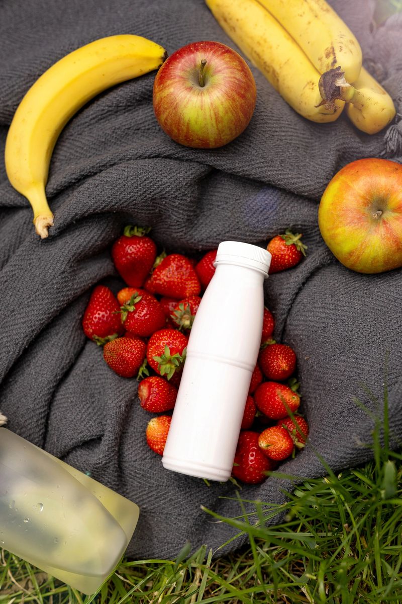 A vibrant picnic scene with fresh fruits, beverages, and a cozy blanket on lush green grass. Three white yogurt bottles without labels, ideal as a mockup for branding. Perfect for promoting healthy