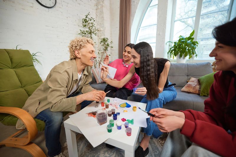 Cheerful friends painting on each other faces with brushes, laughing in cozy living room, focused on creative process. Concept of friendship, fun, creativity, bonding, humor, play, joy, relaxation.