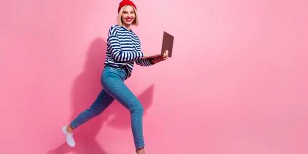 Young woman jumping with laptop against pink backdrop, smiling.