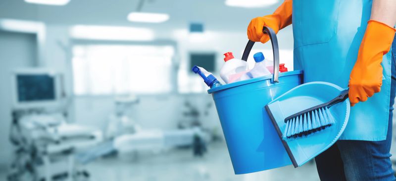 A person wearing orange gloves holds a blue bucket filled with cleaning supplies and a dustpan. The background shows a medical facility with equipment in the distance.