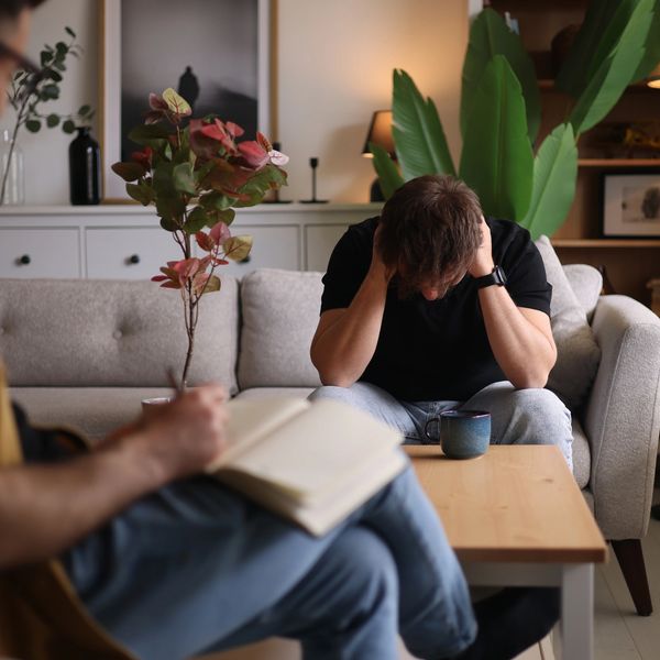 A man sitting on a couch holding his head, another person taking notes.