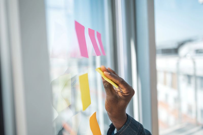 A participant places sticky notes on a glass wall in a bright, professional workspace, emphasizing teamwork, creativity, and organization. This image symbolizes collaboration and the generation of innovative ideas.