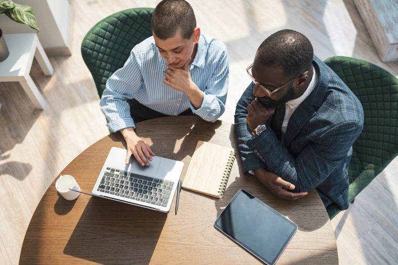 Two professionals, one African American and one Caucasian, discuss ideas while using a laptop in a well-lit office setting. The image represents teamwork, modern technology, and collaborative business efforts.