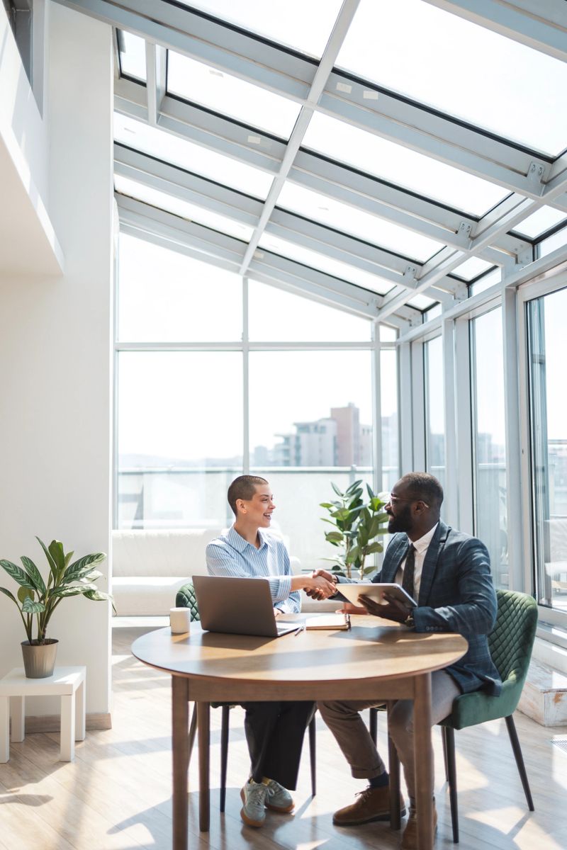 A diverse pair of office workers discuss and collaborate in a bright, natural lit workplace. The environment is modern and professional, emphasizing teamwork, productivity, and positive business relationships.