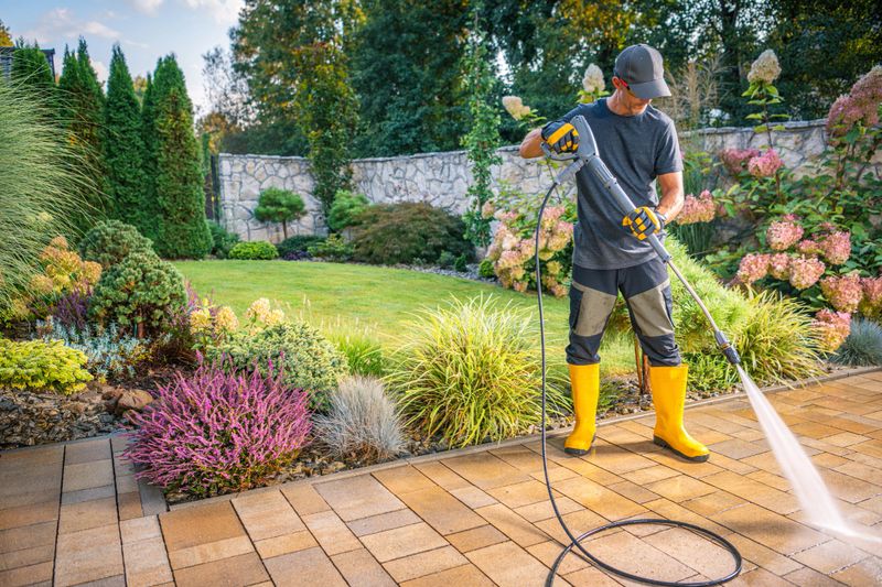 A gardener in bright yellow boots operates a pressure washer on a stone patio, removing dirt and debris. Surrounding colorful flowers and well-maintained shrubbery create a vibrant ambiance.