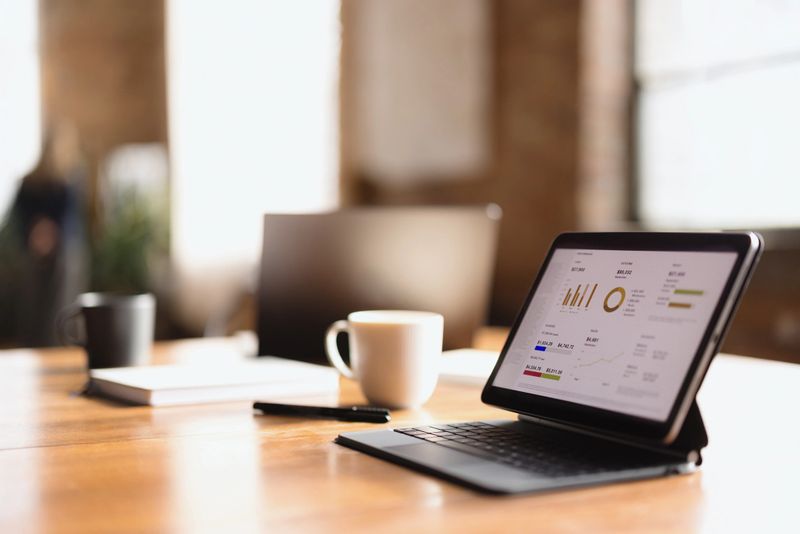 Business data analysis concept with tablet showing charts and graphs, placed on a wooden desk with a cup of coffee and notebook in a modern office setting