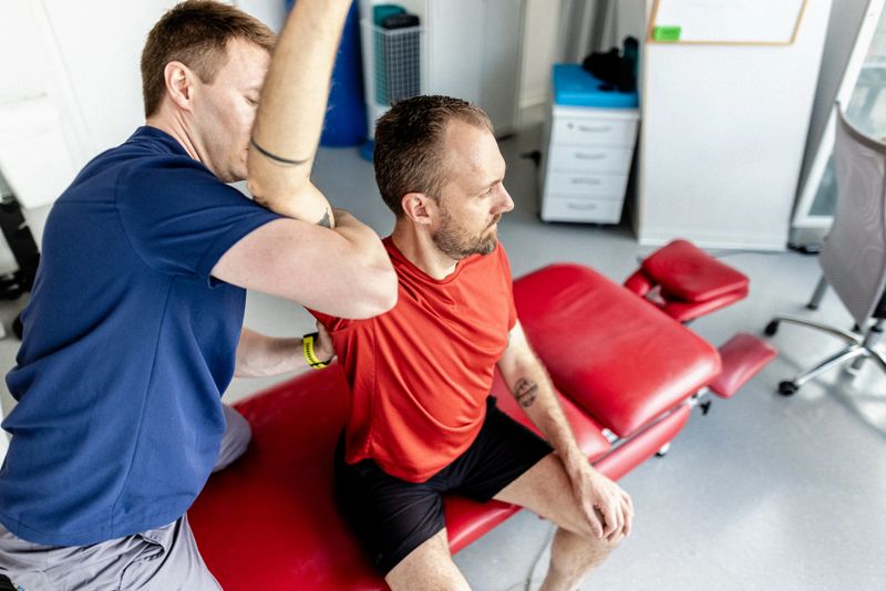 A trained professional providing physical therapy assistance to a patient undergoing shoulder exercises, aiming to restore mobility and alleviate pain, within a modern clinic setting equipped with therapeutic equipment.
