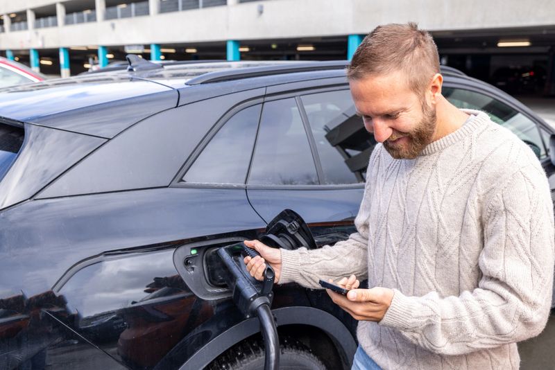 Man with beard uses phone near electric car charging port. Contactless payment technology for EV charging. Modern eco-friendly transportation solutions.