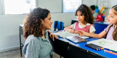 Teacher engaging with two young students at a classroom desk.