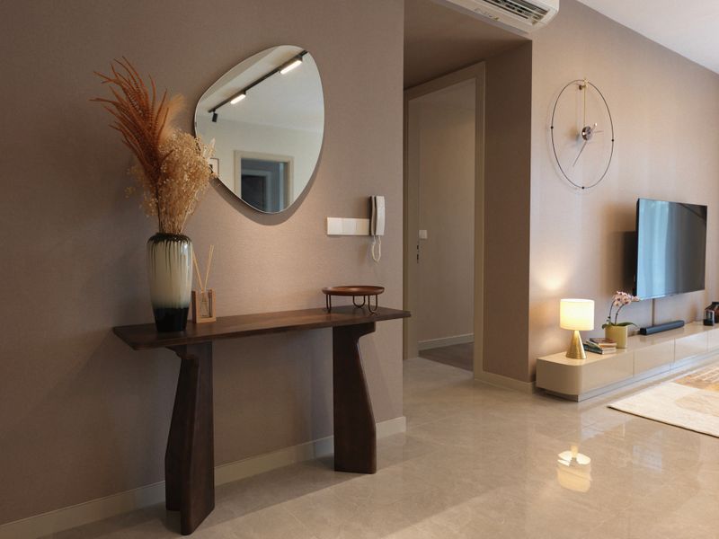 Elegant hallway corner featuring a dark wood console table, decorative vase with dried flowers, and a stylish oval wall mirror. Soft neutral tones, minimalist furniture, and thoughtful lighting design enhance the modern interior. A sleek wall-mounted TV and designer wall clock are visible in the adjacent living room.