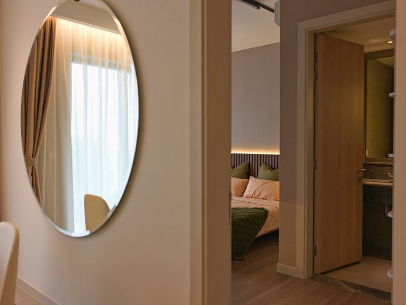 View of a modern bedroom from a hallway featuring a large round wall mirror reflecting daylight. The bedroom is designed with ambient lighting, soft bedding in warm and green tones, and a partially open door leading to a contemporary bathroom.
