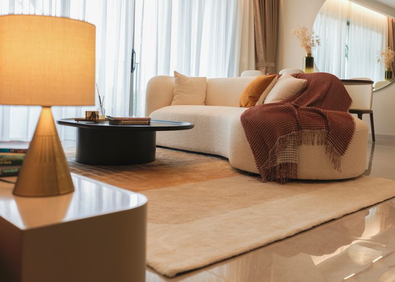 Stylish and minimalistic living room interior featuring a cream-colored curved sofa with neutral cushions and a brown throw blanket. The space is complemented by a round black coffee table, floor-to-ceiling glass doors, and soft natural lighting. Elegant decor includes a large round mirror, indoor plants, and a serene balcony view, creating a warm and inviting atmosphere.