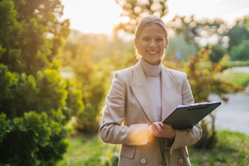 Young businesswoman smiling at the camera while holding a clipboard in a park filled with trees and bushes, basking in the warm glow of sunset during the golden hour
