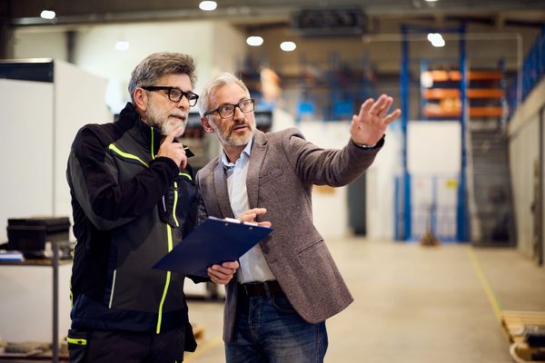 Two men discussing plans inside a spacious industrial warehouse.