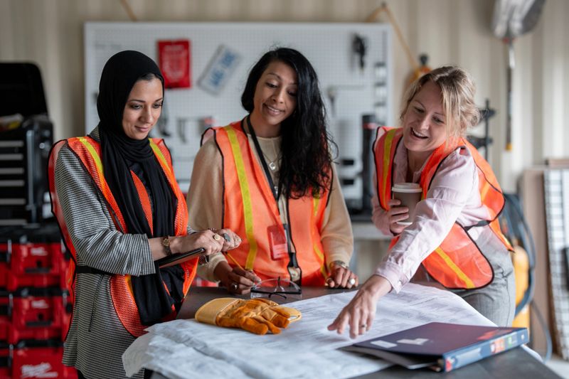 A group of women in safety vests collaborate over construction blueprints in a workshop-like setting, showcasing teamwork, leadership, and skilled roles in construction and architecture.