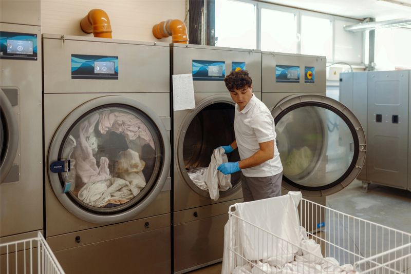 Male laundry worker in protective gloves handles freshly laundered linens in a commercial laundry room equipped with industrial washing machines. The scene emphasizes cleanliness and efficiency in a professional setting.