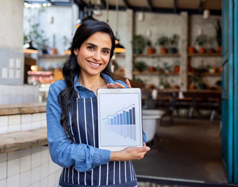 Happy Latin American waitress at a restaurant showing a growth graph on a digital tablet and smiling at the camera