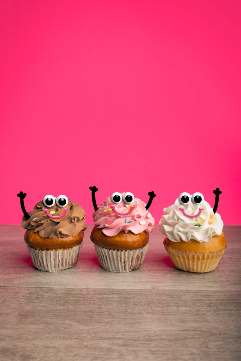Three cute cupcakes with googly eyes and smiles with little arms raised celebrating on a hot pink background.