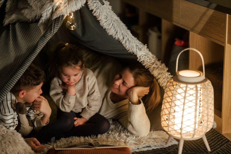 A loving mother spends quality time with her young son and daughter inside a beautifully decorated blanket fort in their bedroom. The soft glow of fairy lights and a woven lantern creates a warm and magical atmosphere as they read a book together.