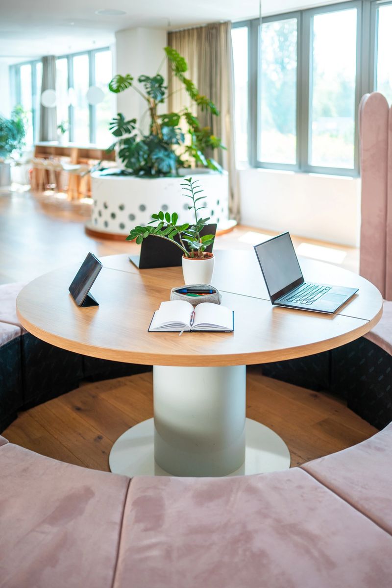 A sleek laptop, notebook, and smartphone rest on an elegant desk, hinting at professional tasks in a stylish, sunlit office with green plants enhancing the serene ambiance.