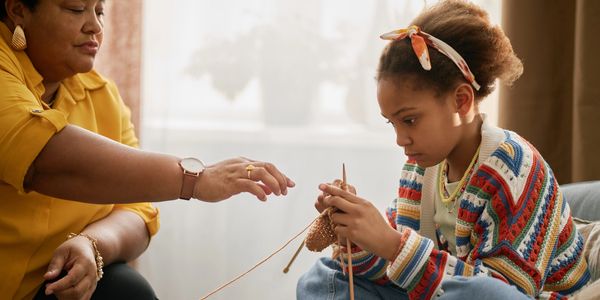A young girl focused on knitting while an older woman guides her.