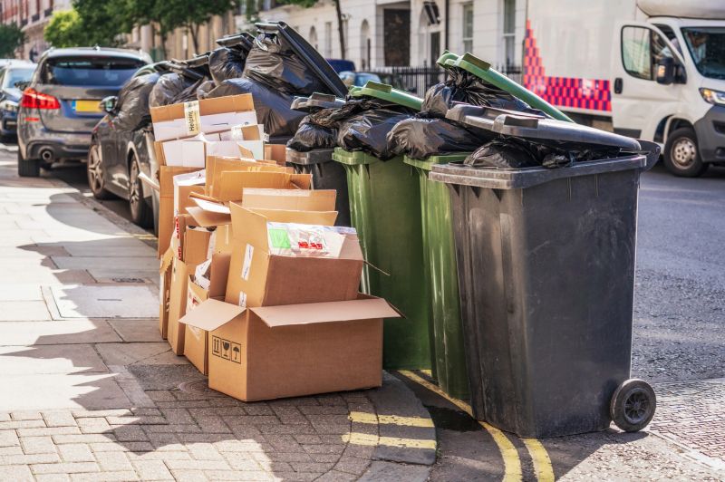 Several black and green wheel bins full of rubbish bags, alongside a collection of cardboard boxes for recycling on a residential street in London.