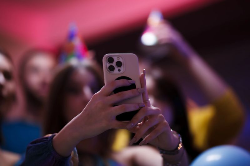 A joyful gathering of friends celebrating a special occasion, embracing the modern trend of capturing moments with smartphones, against a backdrop of colorful lighting and cheerful party decorations.