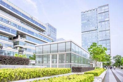 Modern urban buildings with glass facades and green landscaping along a sidewalk.