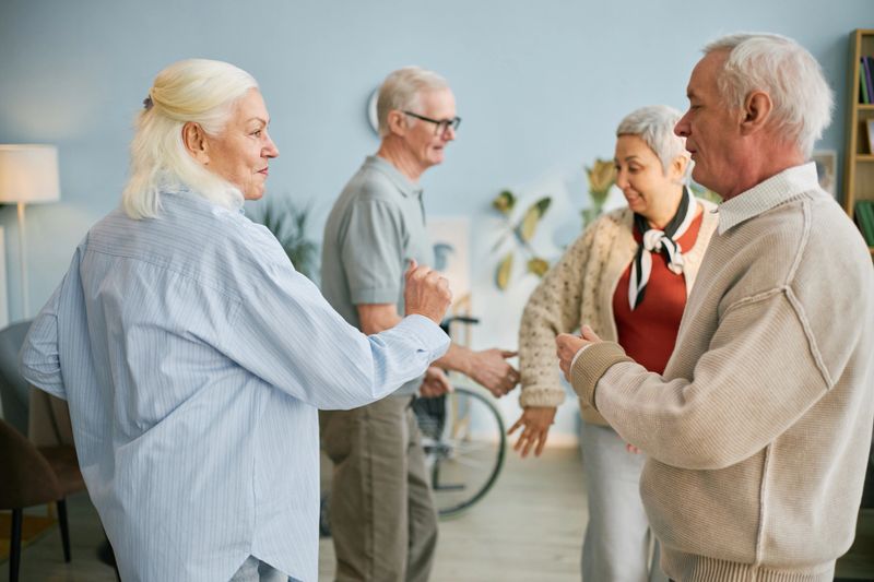 Group of seniors participating in lively conversation in bright living room while standing and smiling at each other, surrounded by cozy furniture and decoration