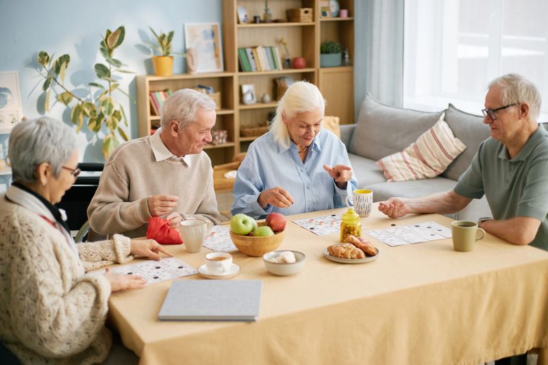 Group of senior friends eating breakfast in retirement home sitting around table, sharing stories and laughter. Table set with fruit and pastries, surrounded by cozy, homely atmosphere