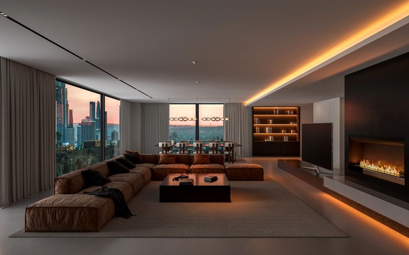 a wide view of the living room in the evening. A large brown sectional sofa, a low coffee table, and a view of the city skyline through large glass walls at dusk dominate the scene. Warm, subdued lighting (LED strips along the ceiling and above the fireplace) creates a cozy atmosphere. In the background, a dining table and a bookshelf are visible. 3D render.