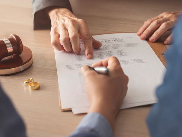 Person signing a divorce agreement with wedding rings and gavel nearby.