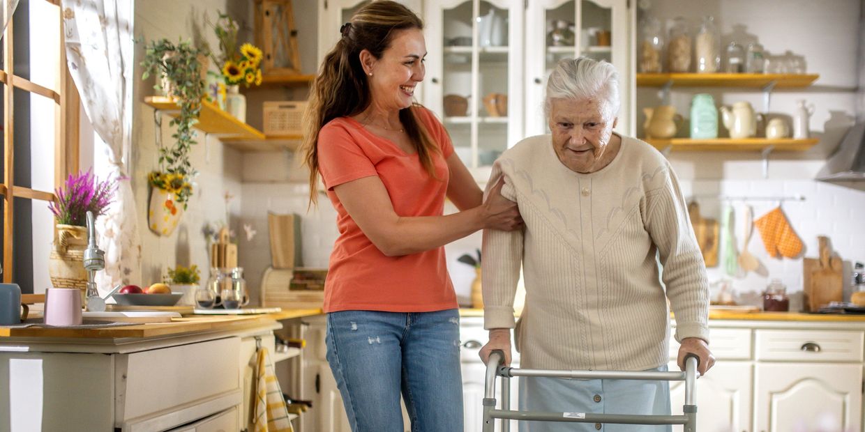 A smiling woman assists an elderly woman using a walker in a cozy kitchen.