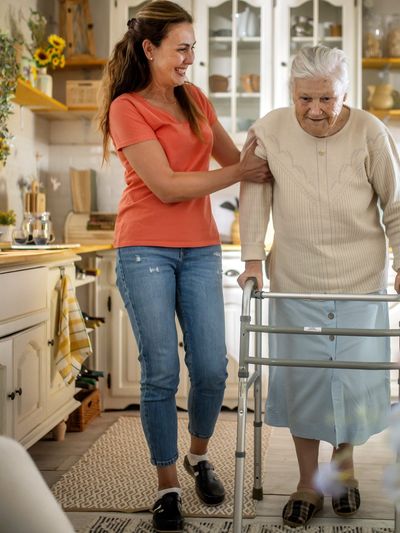 Woman helping elderly woman walk in cozy kitchen.