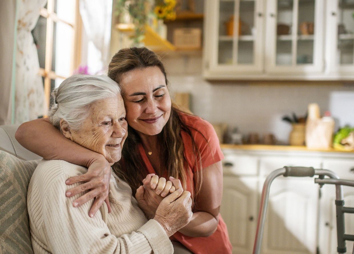 A young woman lovingly embraces an elderly woman sitting on a couch.