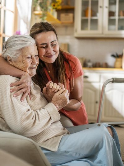 A young woman lovingly embraces an elderly woman sitting on a couch.