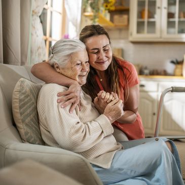 A young woman warmly embraces an elderly woman sitting on a couch.