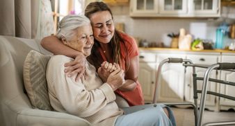 A caregiver warmly embraces an elderly woman sitting in a chair at home.