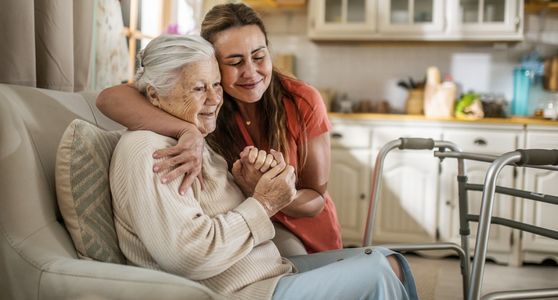 A young woman lovingly embraces an elderly woman sitting on a couch.