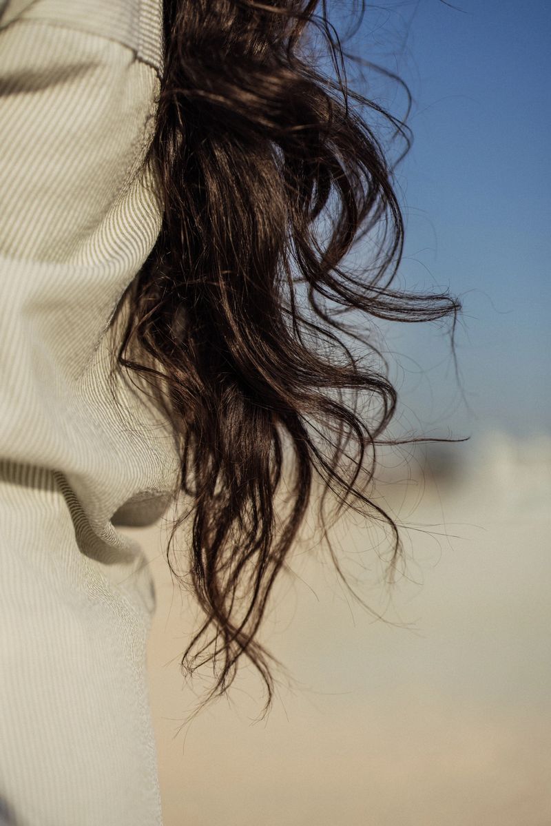 Detailed view emphasizing natural wavy brown hair, softly lit by sunlight. The background features a blurred sandy setting and a clear blue sky, evoking a tranquil outdoor atmosphere. Perfect for highlighting themes of natural beauty and a serene connection with nature.