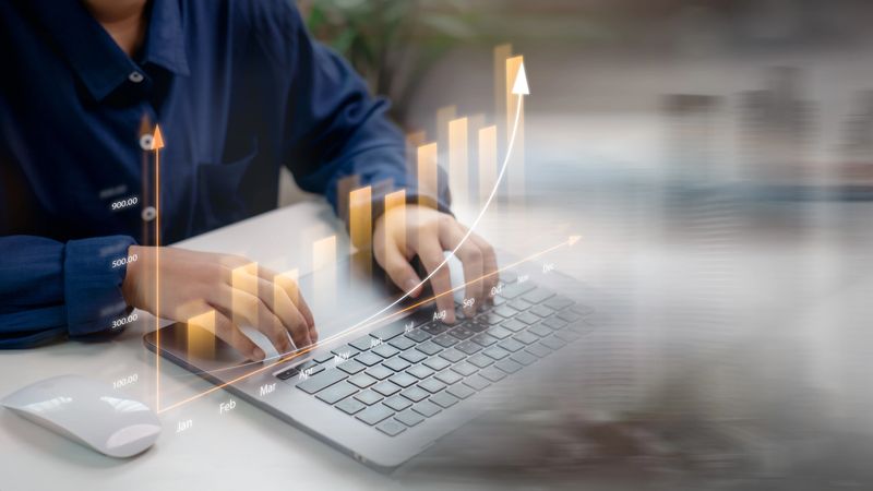Businessperson analyzing financial growth with monthly bar chart and upward trend line on laptop. Concept of business analytics, performance tracking, and data-driven strategy.