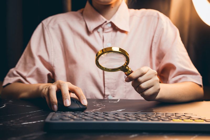 Person Holding Magnifying Glass While Using Computer Mouse in Office Environment for Investigation or Analysis