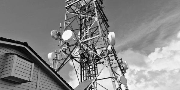 Black and white image of a communication tower next to a building under a cloudy sky.