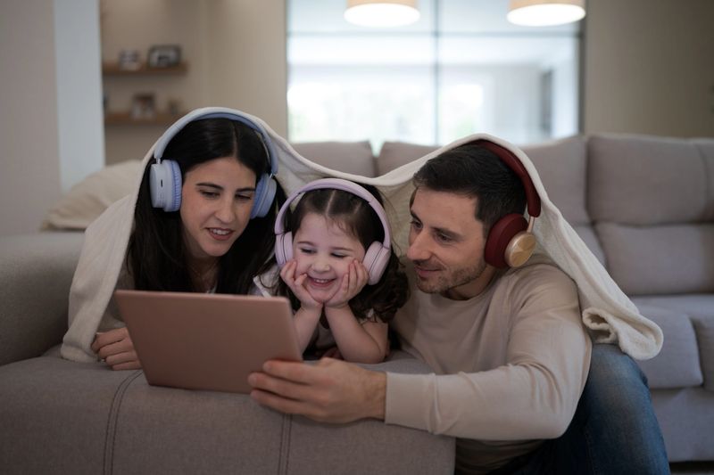 Happy family composed by mother, father and daughter enjoying time together watching a movie on a tablet under a blanket with headphones on