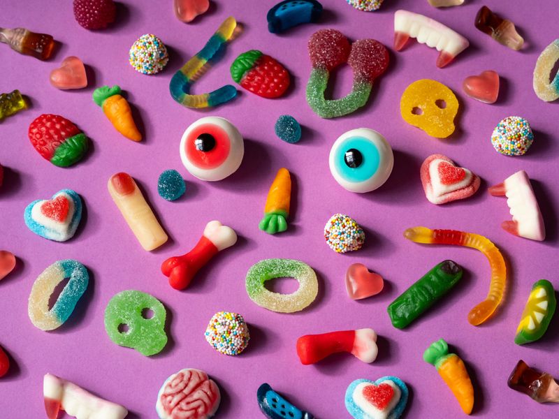 An array of vibrant gummy candies arranged on a purple background showcases a face and various shapes and colors, creating a playful and cheerful display perfect for any sweet tooth.