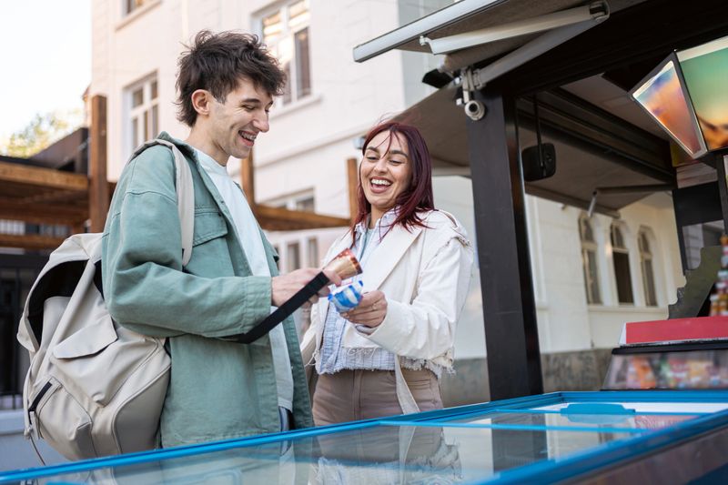 A cheerful man and woman enjoy selecting ice cream from an outdoor stand on a pleasant day, showcasing friendships and joyful moments in an urban setting.
