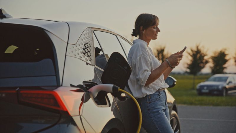 A mid adult woman stands near a charging electric car in a parking lot at sunset, using her smartphone. She wears a white shirt and blue jeans, with a tranquil background featuring trees and parked cars.