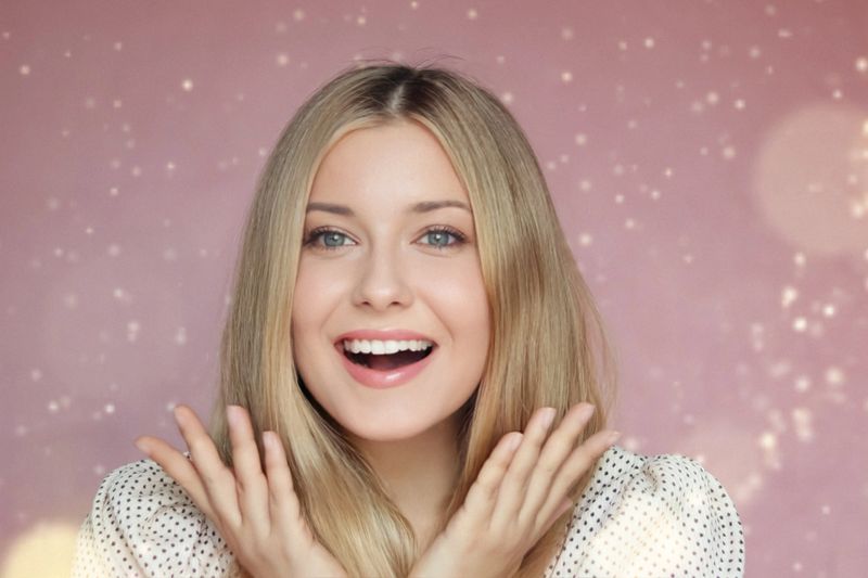 Holiday joy and natural beauty, smiling blonde woman with blue eyes expressing surprise and excitement against a sparkling pink festive background