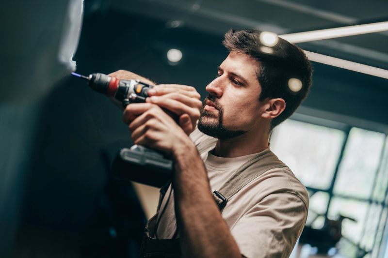 A skilled craftsman focuses on his work, using a power drill to attach materials in a bright, modern workshop filled with tools and natural light.