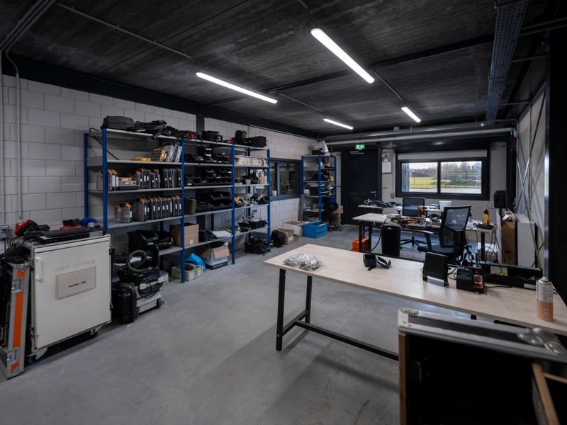 Clean, organized workshop featuring metal shelving units stocked with tools and supplies along white tiled walls. Concrete floors and exposed ceiling with LED strip lighting. Multiple workbenches, storage cabinets, and a window providing natural light create an efficient workspace for DIY projects and professional craftsmanship.